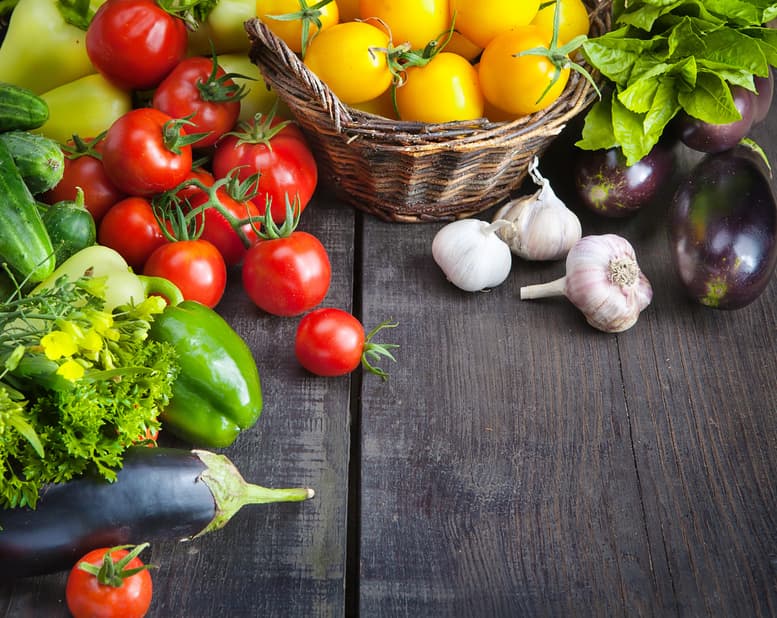Colorful assortment of fresh vegetables including tomatoes, peppers, eggplants, garlic, and leafy greens, arranged on a wooden surface, emphasizing healthy eating and nutrition during winter months.
