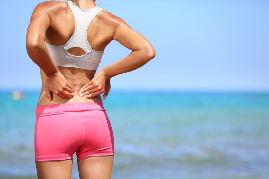 Woman in sports attire experiencing lower back pain at the beach, representing common issues treated by chiropractors like back pain and muscle tension.