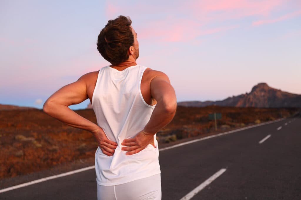 Man in a sleeveless shirt experiencing back pain while standing on a road, emphasizing the importance of proper lifting techniques to avoid spring injuries.