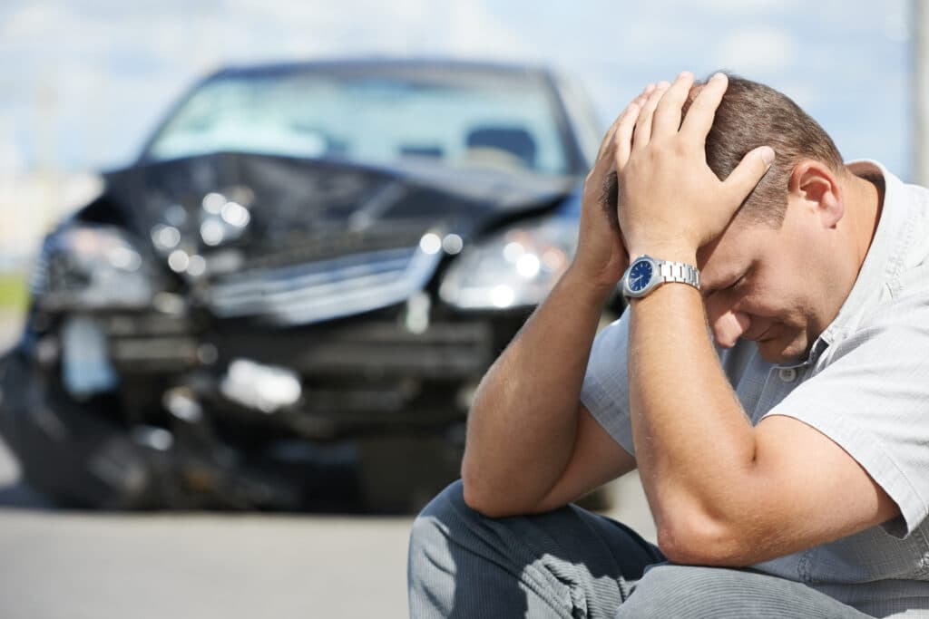 Man sitting on the ground with his head in his hands, looking distressed after a car accident, with a damaged black car in the background, illustrating the emotional impact of auto accidents and the importance of chiropractic care for recovery.