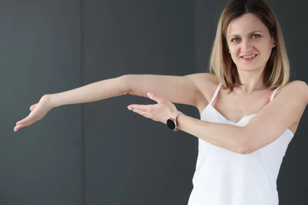 Woman demonstrating arm extension, illustrating hypermobility, wearing a white tank top, smiling, against a neutral background.